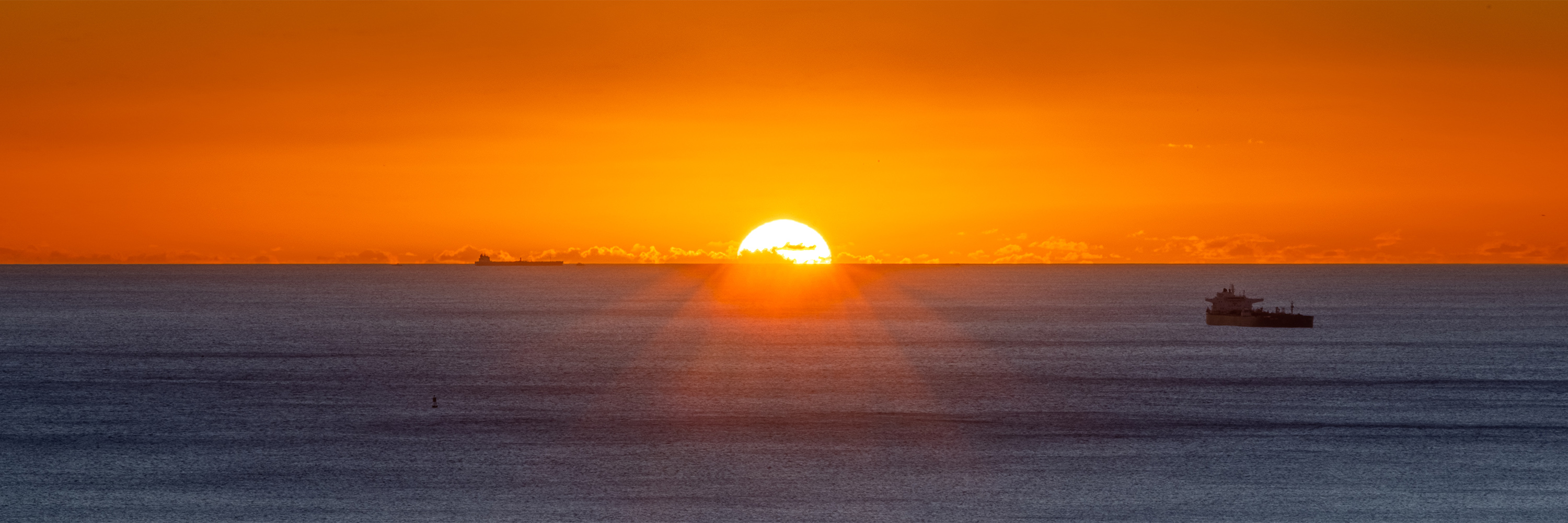 A stunning sunset off the coast of Honolulu dominated by the orange sky and blue ocean, punctuated by shipping boats.